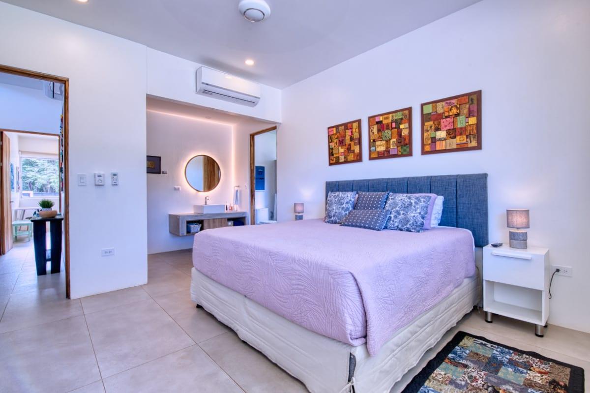 The main bedroom with a king bed, upholstered headboard and three framed artworks above it, opening to an ensuite with a round backlit mirror