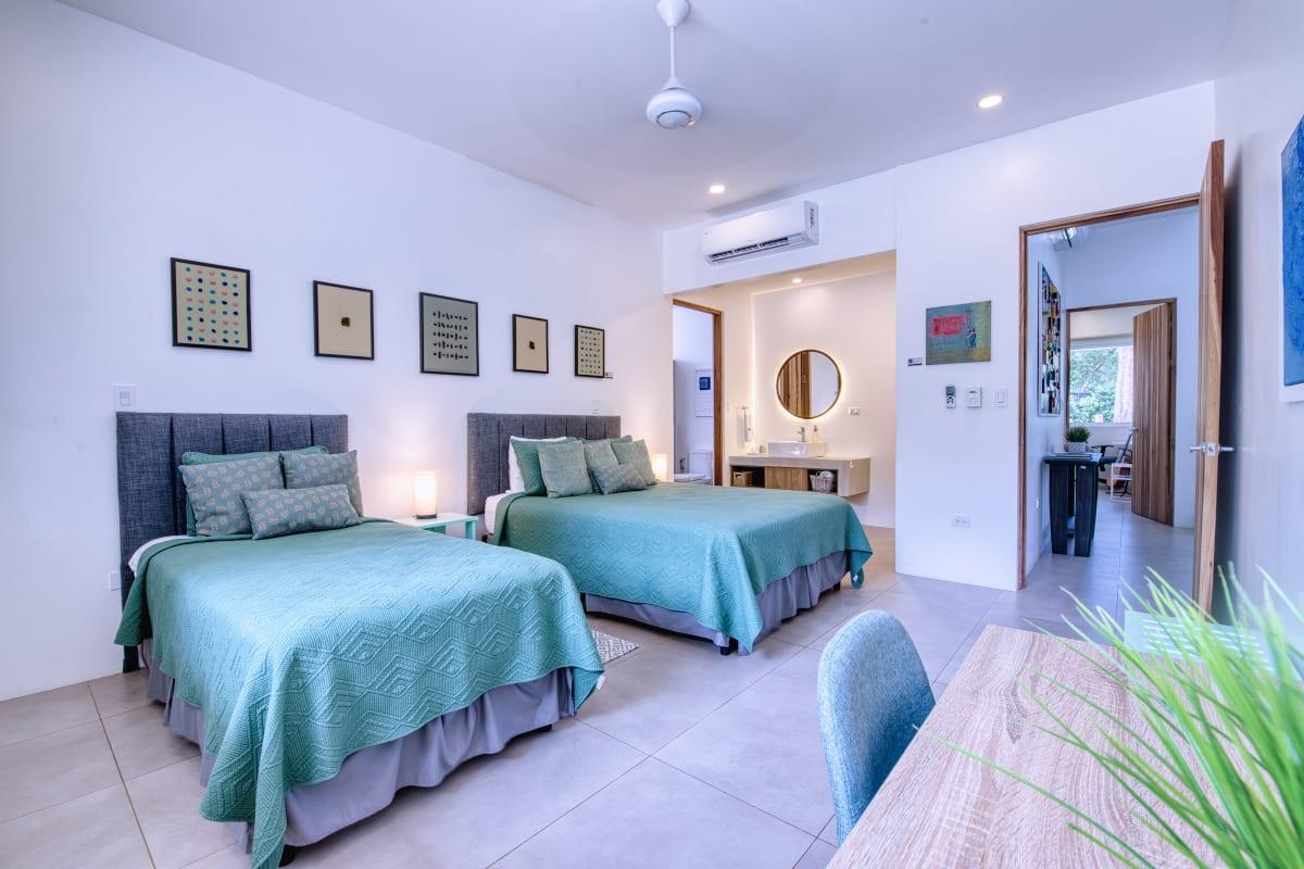 The second bedroom with two beds dressed in teal, a row of small framed works on the wall and an ensuite beyond