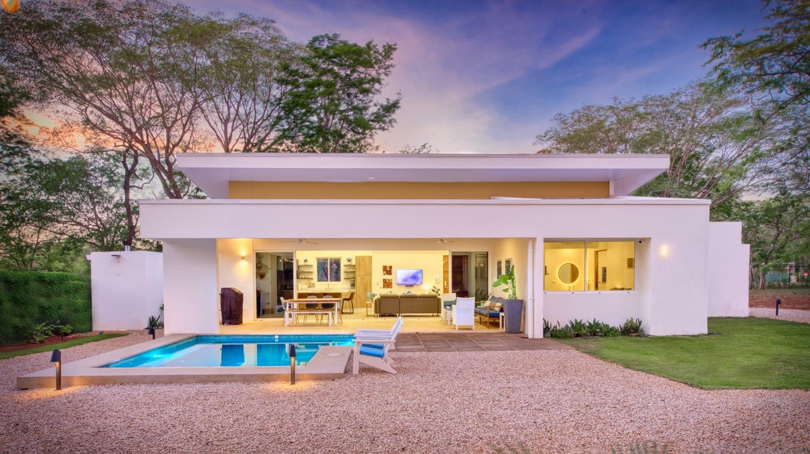 The house at dusk seen head-on, warm light spilling from the open living space onto the lit pool