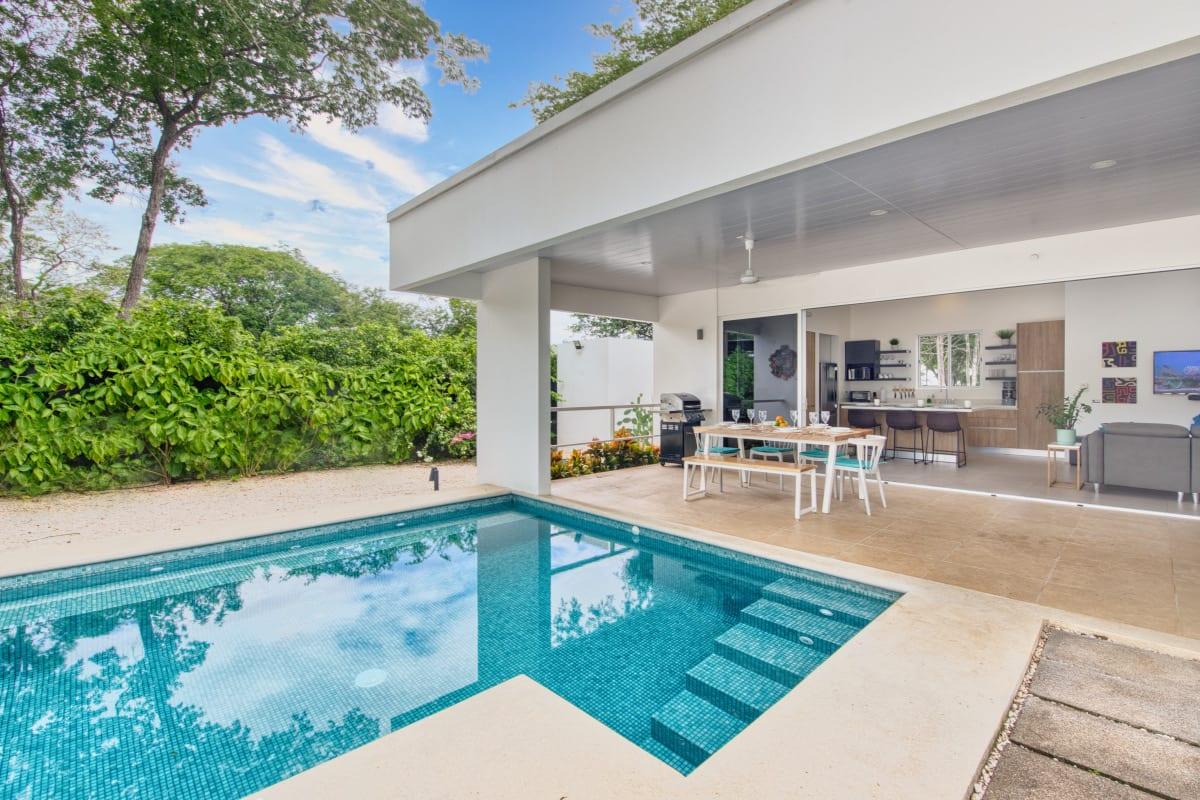 The turquoise mosaic pool in daylight, with the covered terrace, dining table and kitchen visible beyond