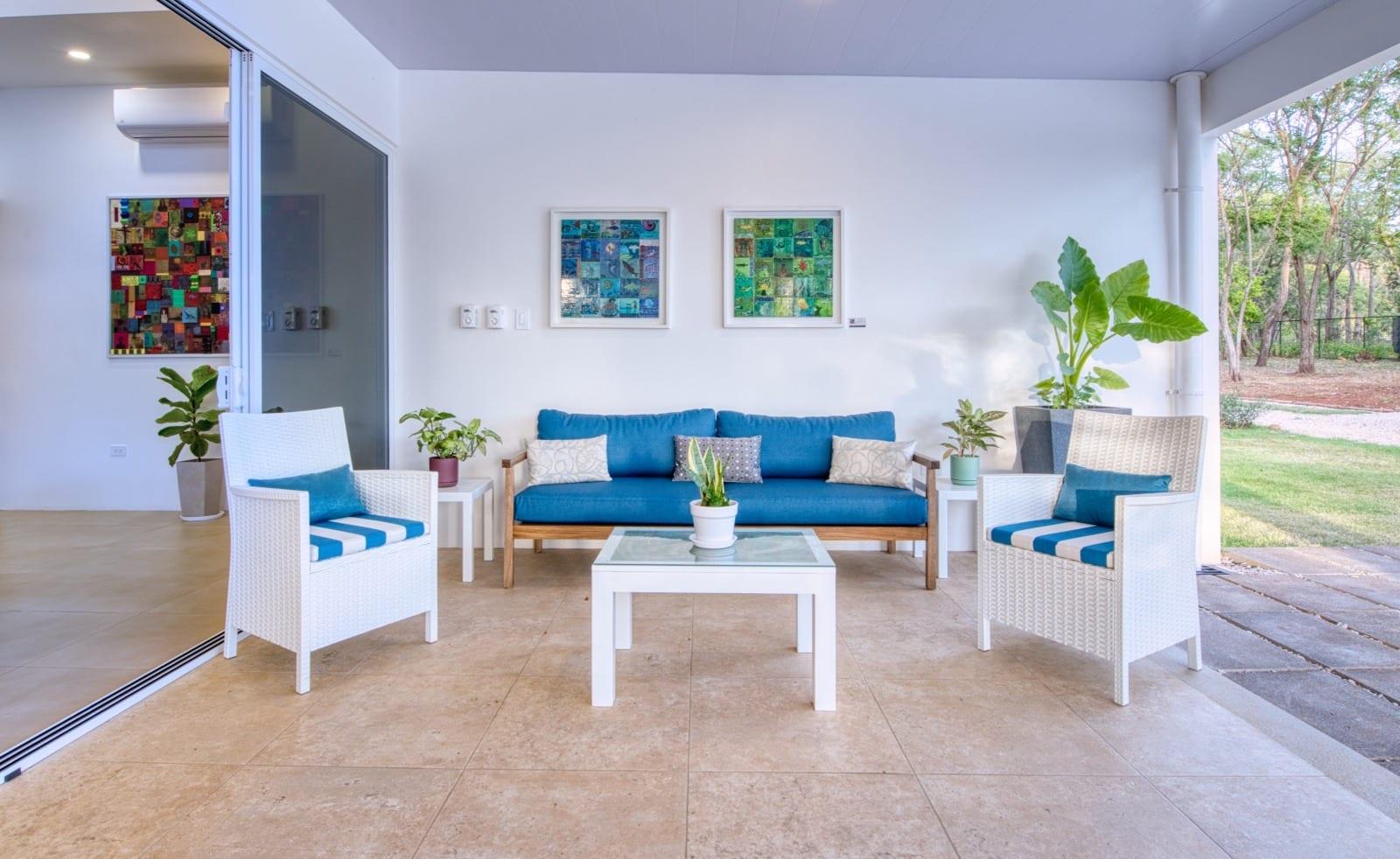The covered terrace lounge, two framed works on the white wall above a blue sofa, the garden beyond
