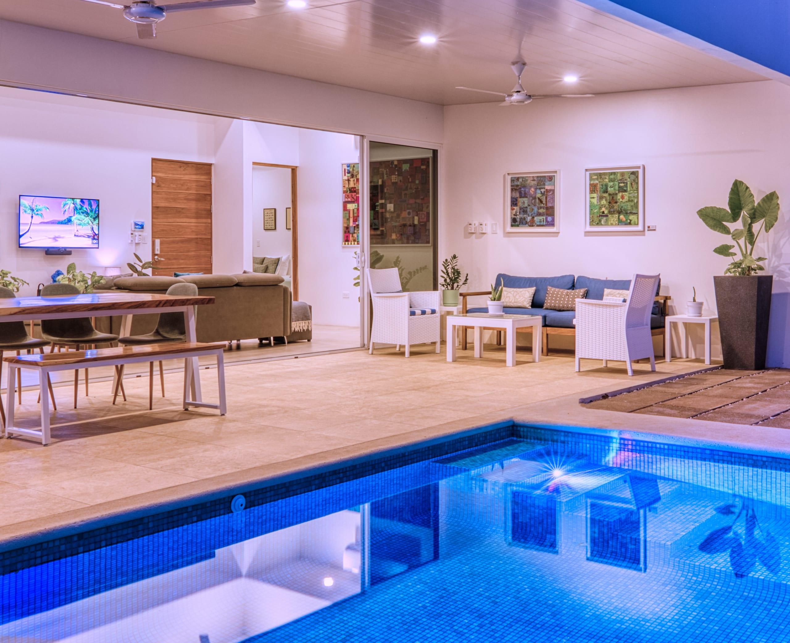 The terrace at night, the pool lit blue, with lounge seating, a long wooden dining table and framed artworks on the wall